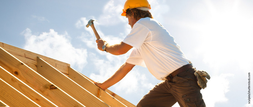 Dachdecker mit gelbem Schutzhelm beim Befestigen von Konstruktionsvollholz (KVH) auf einem Dachstuhl unter strahlendem Himmel.