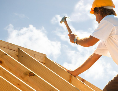 Dachdecker mit gelbem Schutzhelm beim Befestigen von Konstruktionsvollholz (KVH) auf einem Dachstuhl unter strahlendem Himmel.