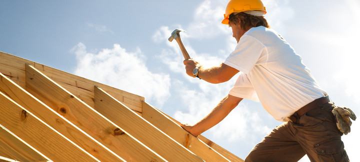 Dachdecker mit gelbem Schutzhelm beim Befestigen von Konstruktionsvollholz (KVH) auf einem Dachstuhl unter strahlendem Himmel.